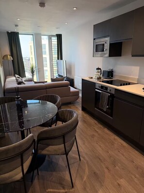 A modern kitchen with a microwave, oven, and a dining area with a table and chairs. - Apartment 1 Bedroom (Salford)