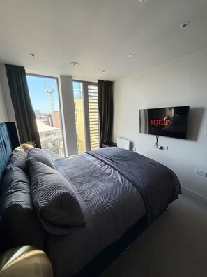 Ein modernes Schlafzimmer mit einem großen Bett, einem an der Wand befestigten Fernseher und Blick auf die Stadtlandschaft durch das Fenster.