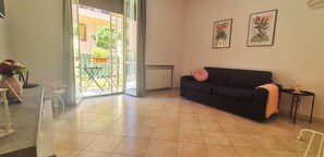 Ein Wohnzimmer mit schwarzem Sofa, einem kleinen Tisch mit Lampe und einer Schiebetür zu einem Außenbereich mit Tisch und Stühlen.