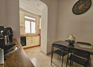 A dining area with a table, chairs, a clock, and a view into a kitchen with cabinets and a window. - Casalù | Private Garden, Free Parking & Metro B to Downtown Rome (Roma)