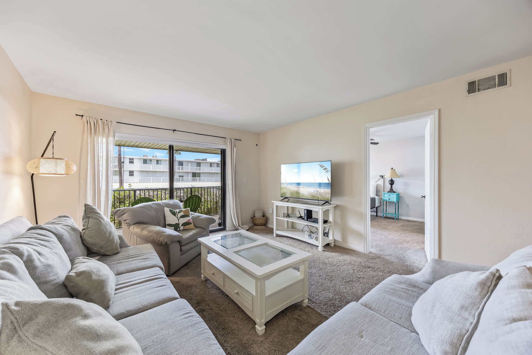 A living room with a sofa, a coffee table, and a TV. There is a balcony with a view of buildings.