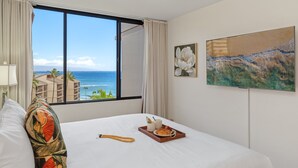 Una camera d'albergo con un letto, una finestra vista oceano, un quadro al muro e un vassoio con prodotti per la colazione.