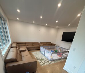 A living room with a sectional sofa, a glass playpen, and a mounted TV. - Luxury 8BR Estate Heated Pool Walk to Shuls (Long Branch)