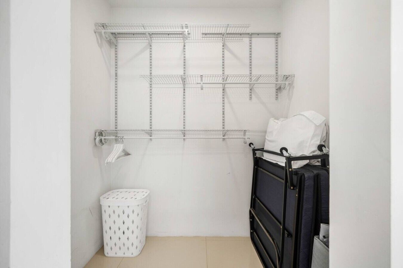 Una habitación pequeña y limpia con una estantería metálica, un cesto de ropa blanca y una estructura de cama negra.