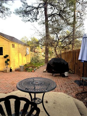 A patio area with a round metal table and chairs, a barbecue grill, and a yellow building in the background. - This house is a 3 bedroom(s), 1 bathrooms, located in Prescott, AZ. (Prescott)