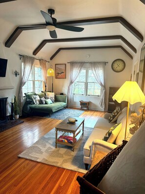 A spacious living room with a ceiling fan, a fireplace, a green sofa, a yellow lamp, and a large window with curtains. - This house is a 3 bedroom(s), 1 bathrooms, located in Prescott, AZ. (Prescott)