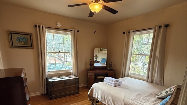Um quarto com cama, penteadeira, cômoda, ventilador de teto e janela com cortinas.