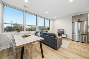 A modern kitchen with a large stainless steel refrigerator, a dining area with a wooden table and chairs, and a grey sofa.