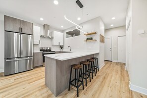 Uma cozinha moderna com geladeira de aço inoxidável, uma ilha branca com banquetas e piso de madeira.