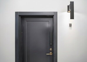 A black door with a keyhole and a lock, a small light fixture above it, and a sign with the number "1B" on the wall.