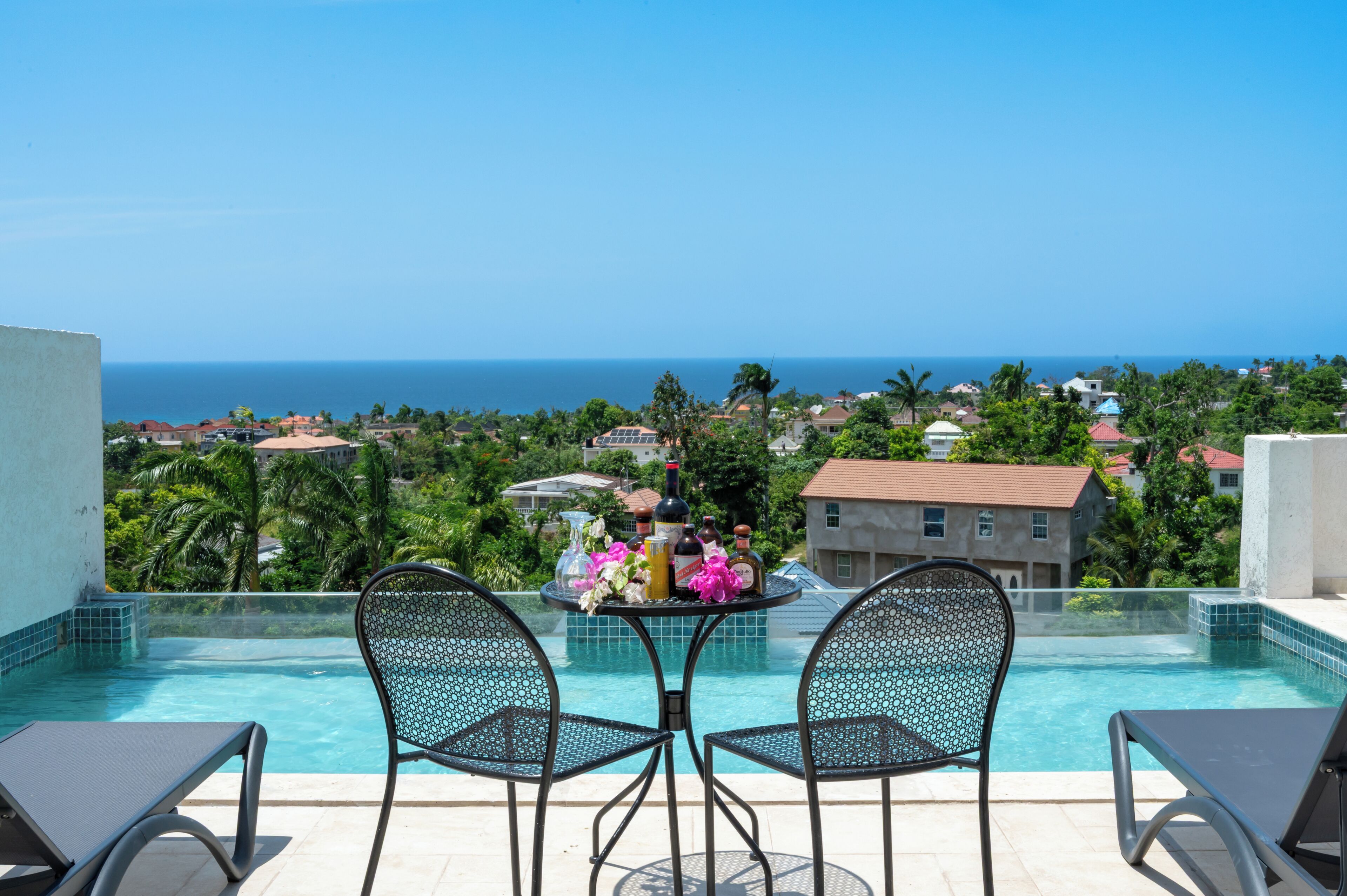 A poolside area with two chairs and a table, overlooking a coastal view with houses and palm trees.