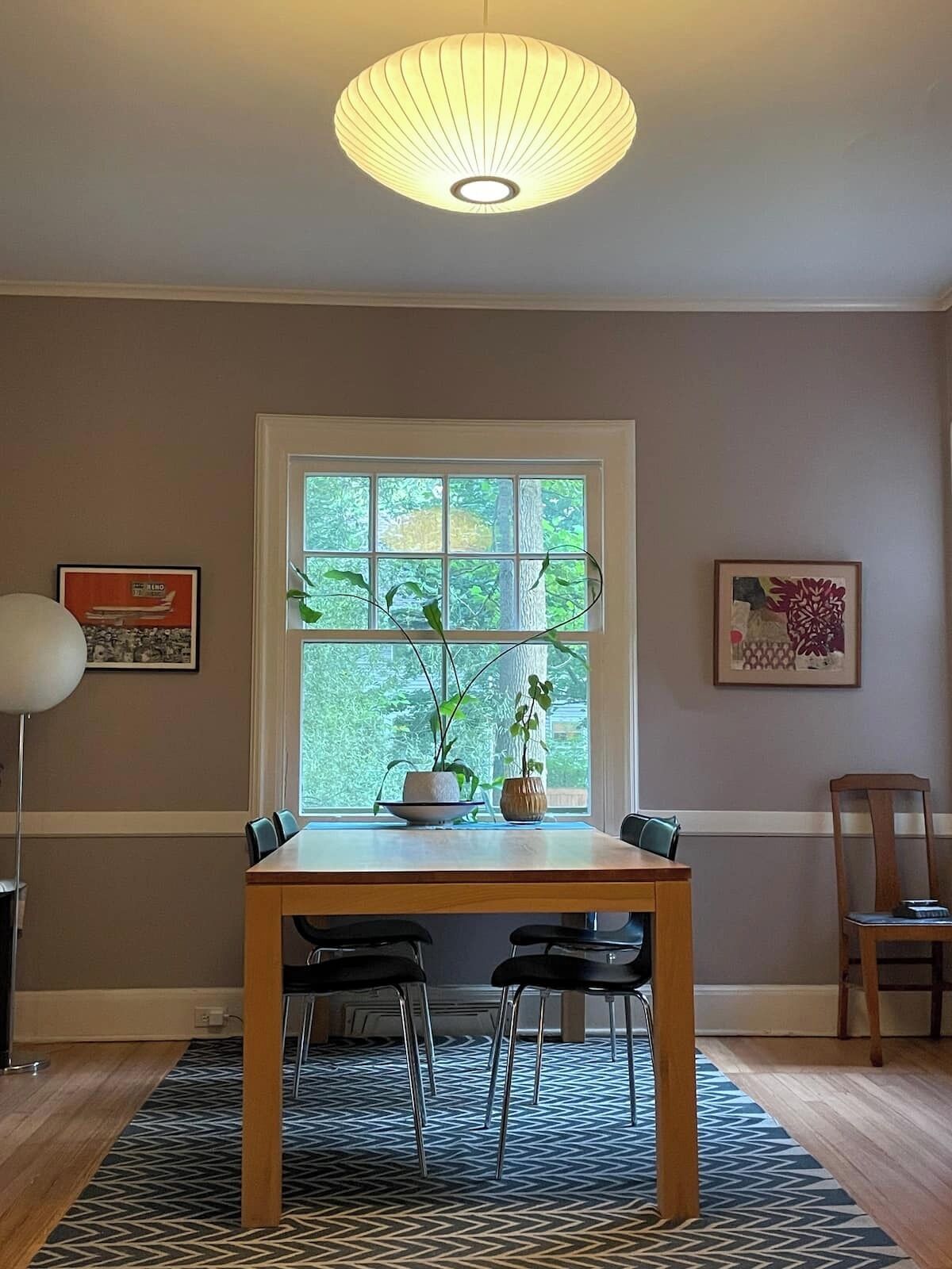 Uma sala de jantar com uma mesa, cadeiras, uma janela e duas plantas em vasos.
