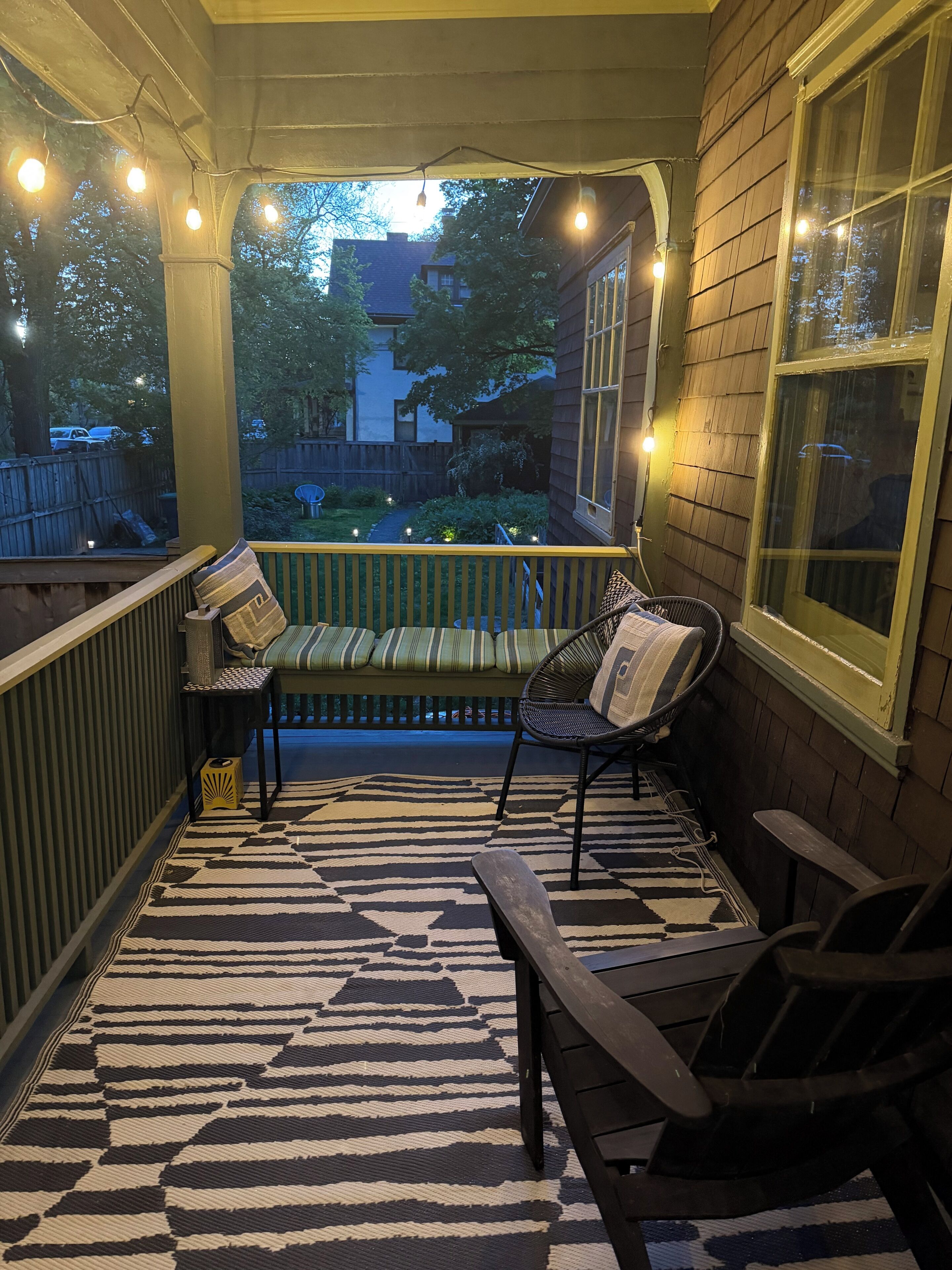 A covered porch with a bench and chairs, a patterned rug, and string lights.