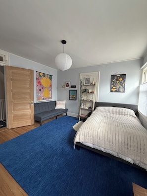 Un dormitorio con cama, un tapete azul, un sofá gris, una lámpara colgante blanca y una pared con cuadros enmarcados.