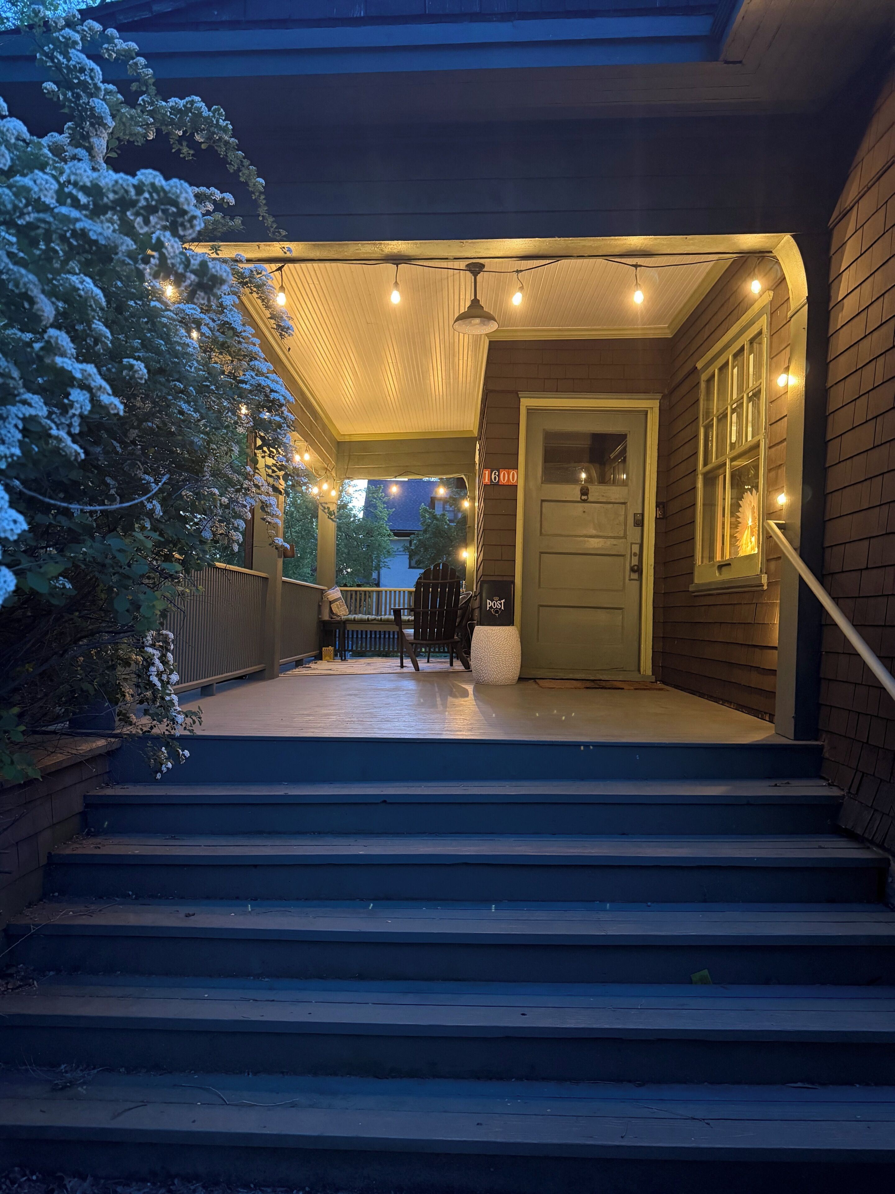A covered porch with steps leading to a door, string lights, and a chair.