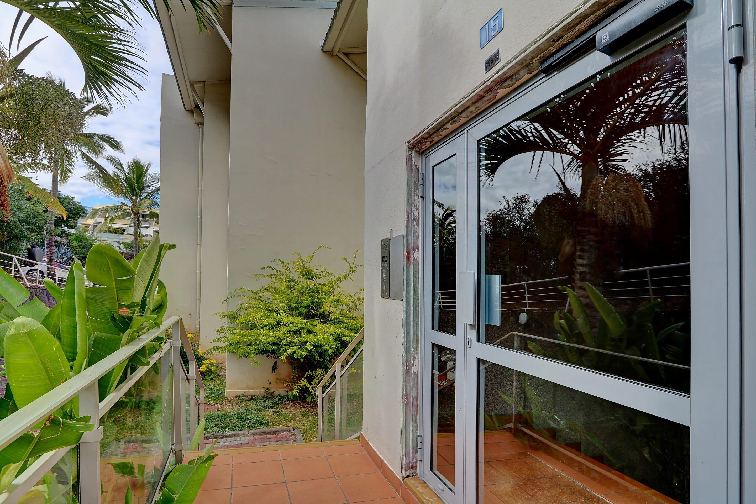 Una entrada de edificio con puerta de vidrio, portón metálico y un pequeño jardín con plantas y una palmera.