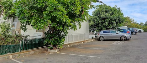 A parking lot with cars, a tree, and a building with a white wall and a green fence.