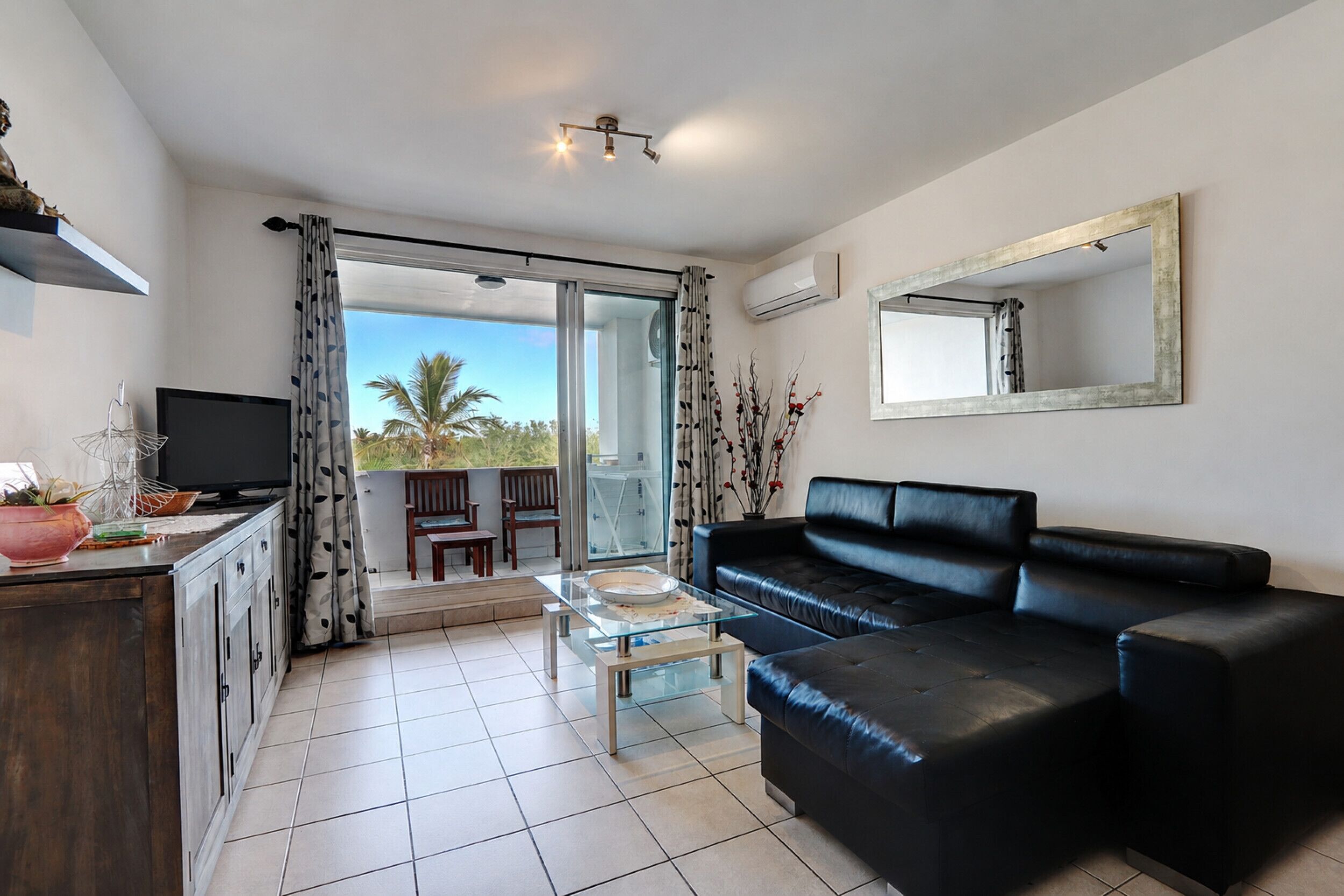 A modern living room with a black leather sofa, a glass coffee table, and a flat-screen TV. There is a balcony with a view of palm trees.
