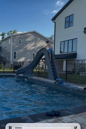 Une piscine avec un toboggan, une maison en arrière-plan et un ciel dégagé.