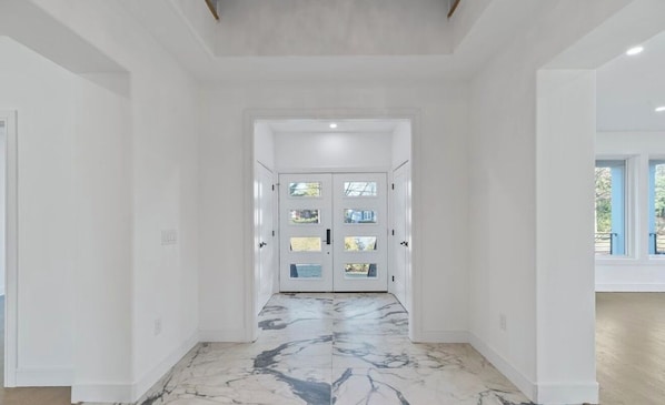 Un couloir aux murs blancs, doté d’un sol en marbre et d’une double porte avec des panneaux de verre.