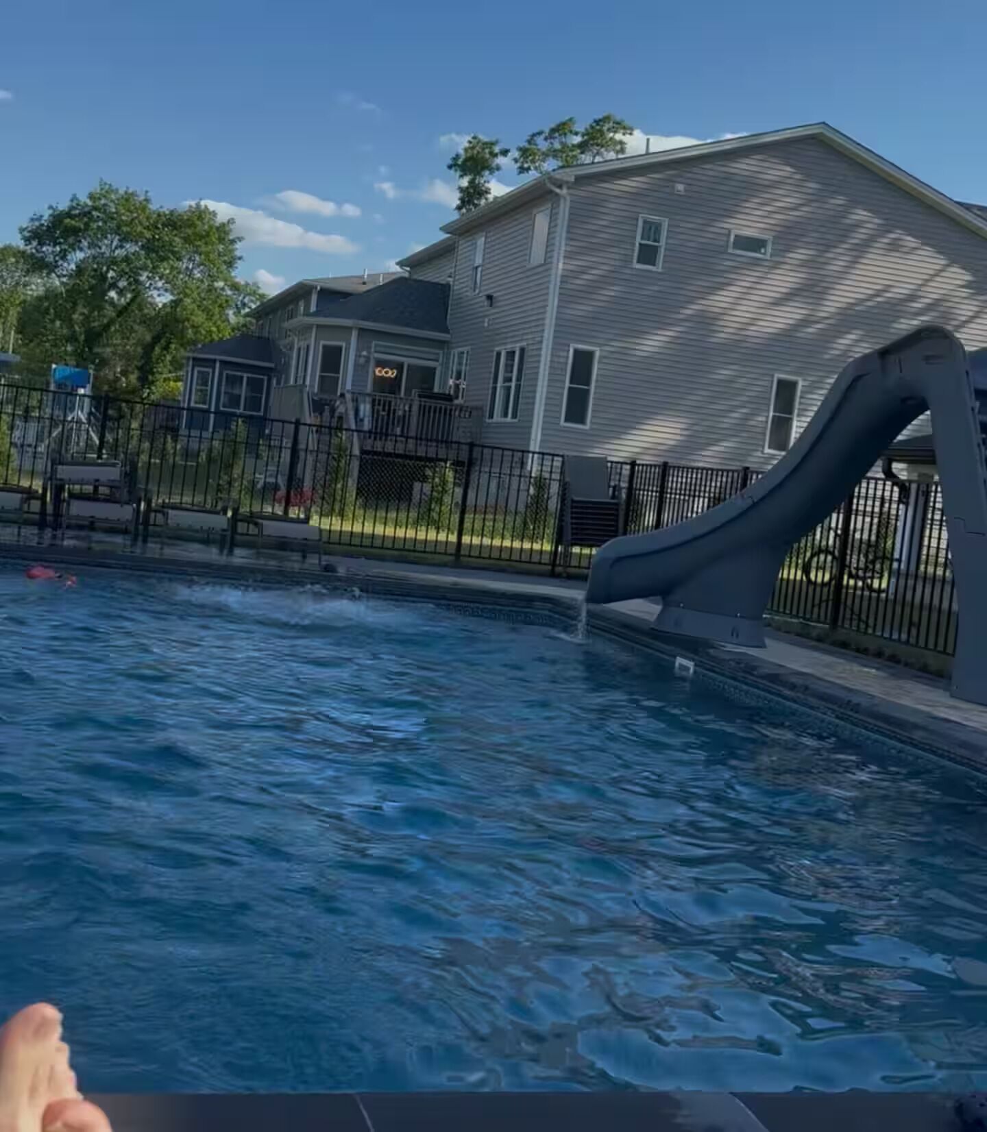 Una piscina con tobogán, rodeada por una cerca, y una casa al fondo.