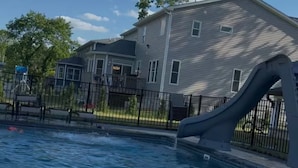 A swimming pool with a slide, surrounded by a fence, and a house in the background.