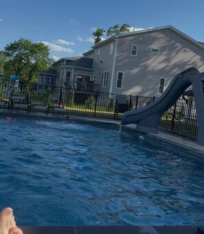 A swimming pool with a slide, surrounded by a fence, and a house in the background. - Luxury 8BR Estate - Heated Pool -Walk to Shuls (Long Branch)