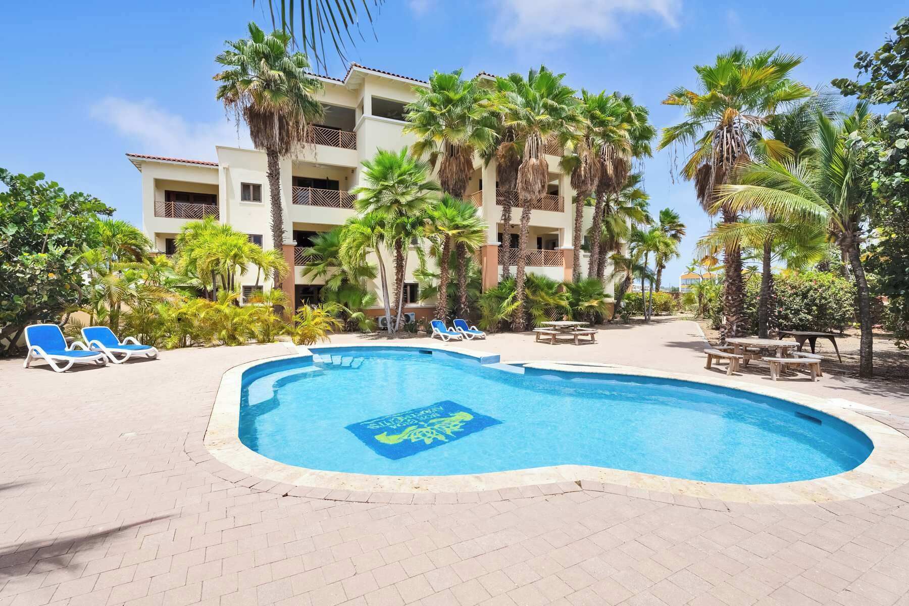 Une piscine entourée de chaises longues et de palmiers, le tout bordé d’un immeuble doté de balcons.