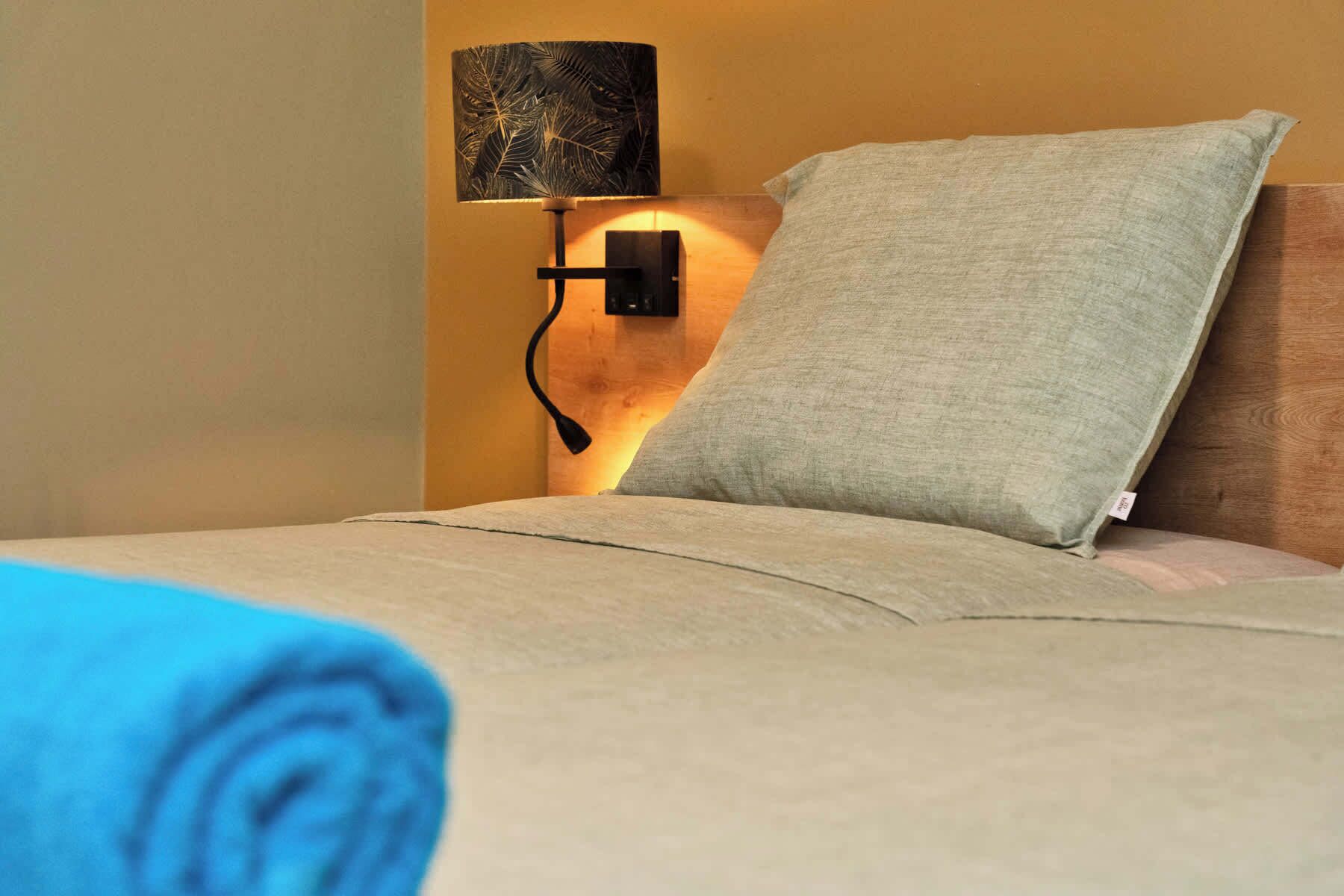 Un lit bien fait, avec un oreiller, une lampe fixée au mur et une serviette enroulée.