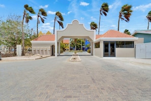 A gated entrance with a central archway, palm trees, and a paved area. - Kas Kurá, garden terrace and pool in Kralendijk (Kralendijk)