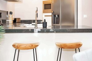 Une cuisine moderne équipée d’un réfrigérateur en acier inoxydable, d’une machine à café noire et de deux tabourets de bar en bois.