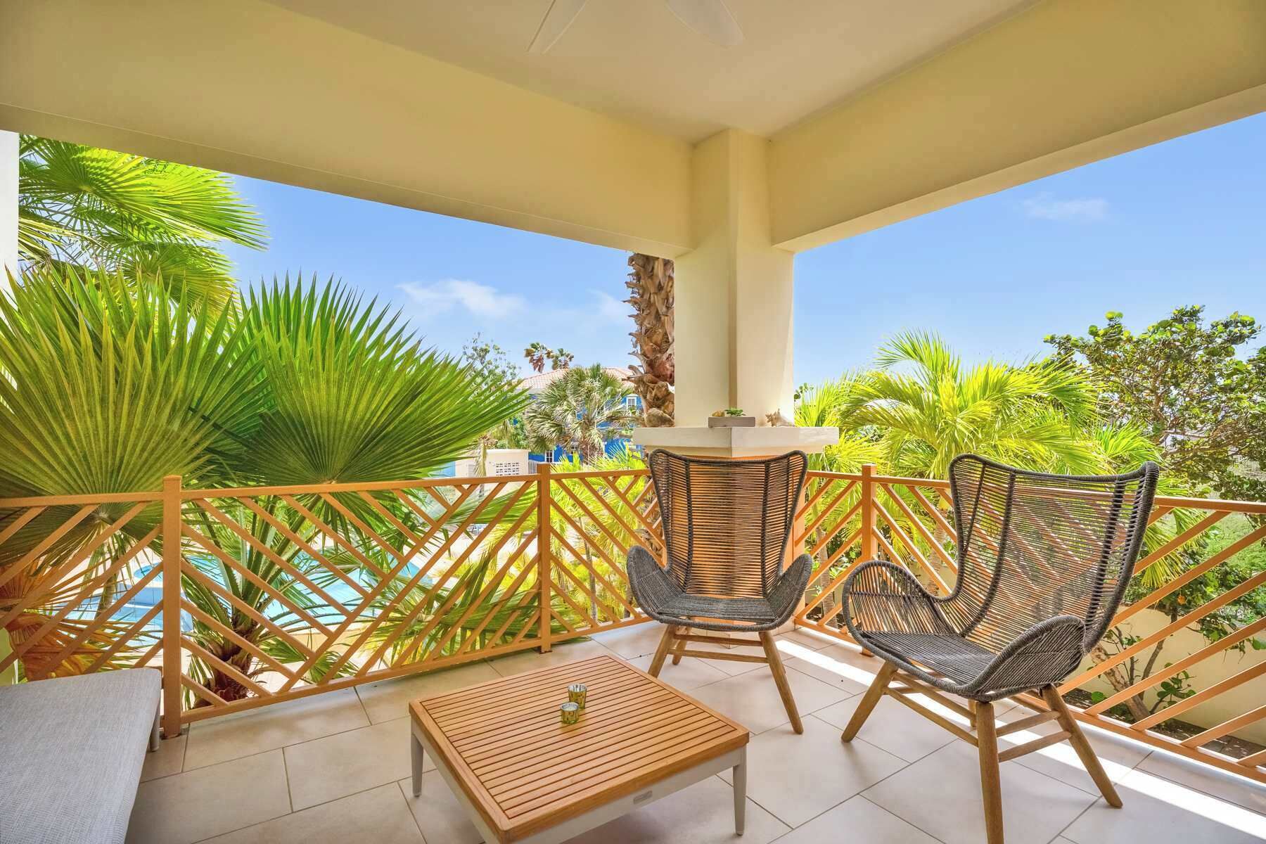 A balcony with two wicker chairs, a wooden table, and a view of palm trees and a building.
