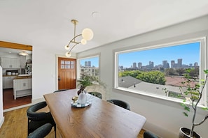 A modern kitchen with white cabinets, a wooden countertop, and a dining area with a wooden table and chairs. A large window offers a view of a city skyline. - Golden Hill Escape Near Balboa Park & Zoo (San Diego)