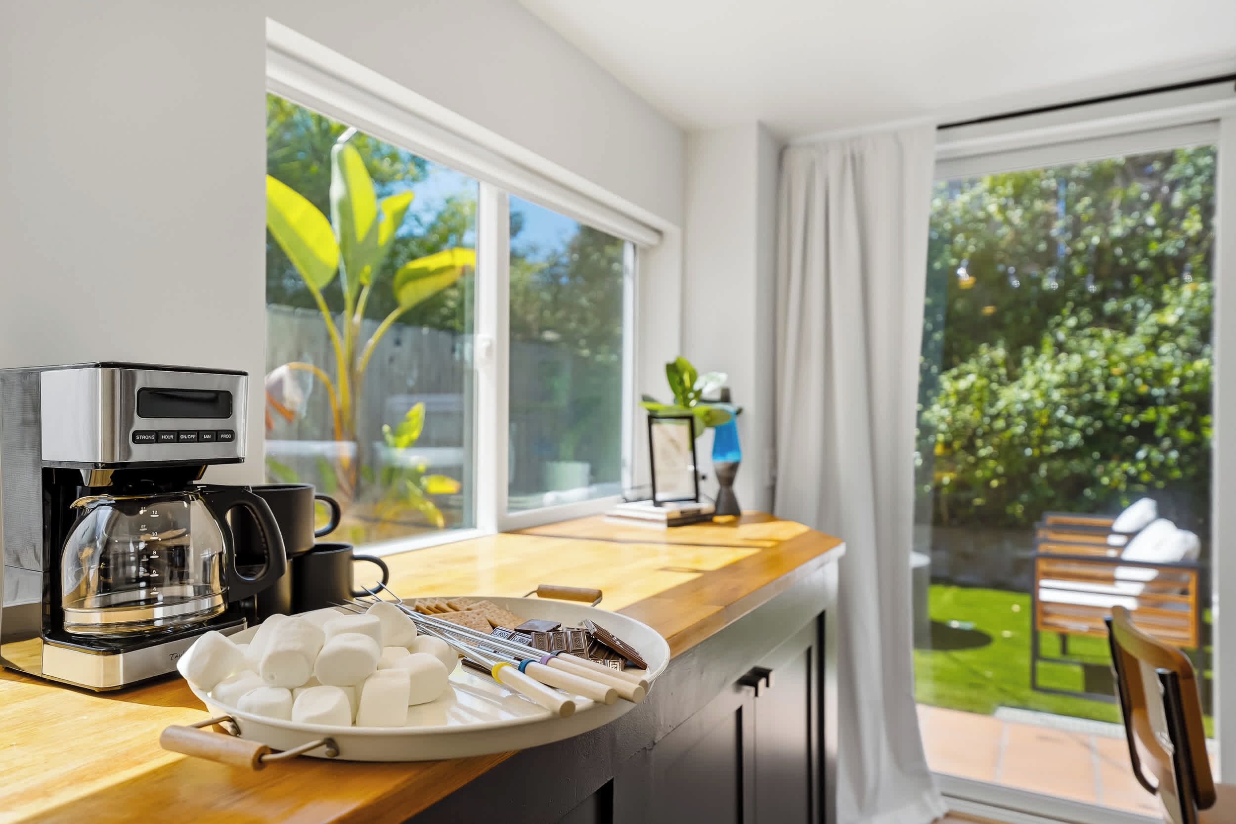 Una cocina con cafetera, un plato de malvaviscos y vistas a un jardín a través de amplios ventanales.