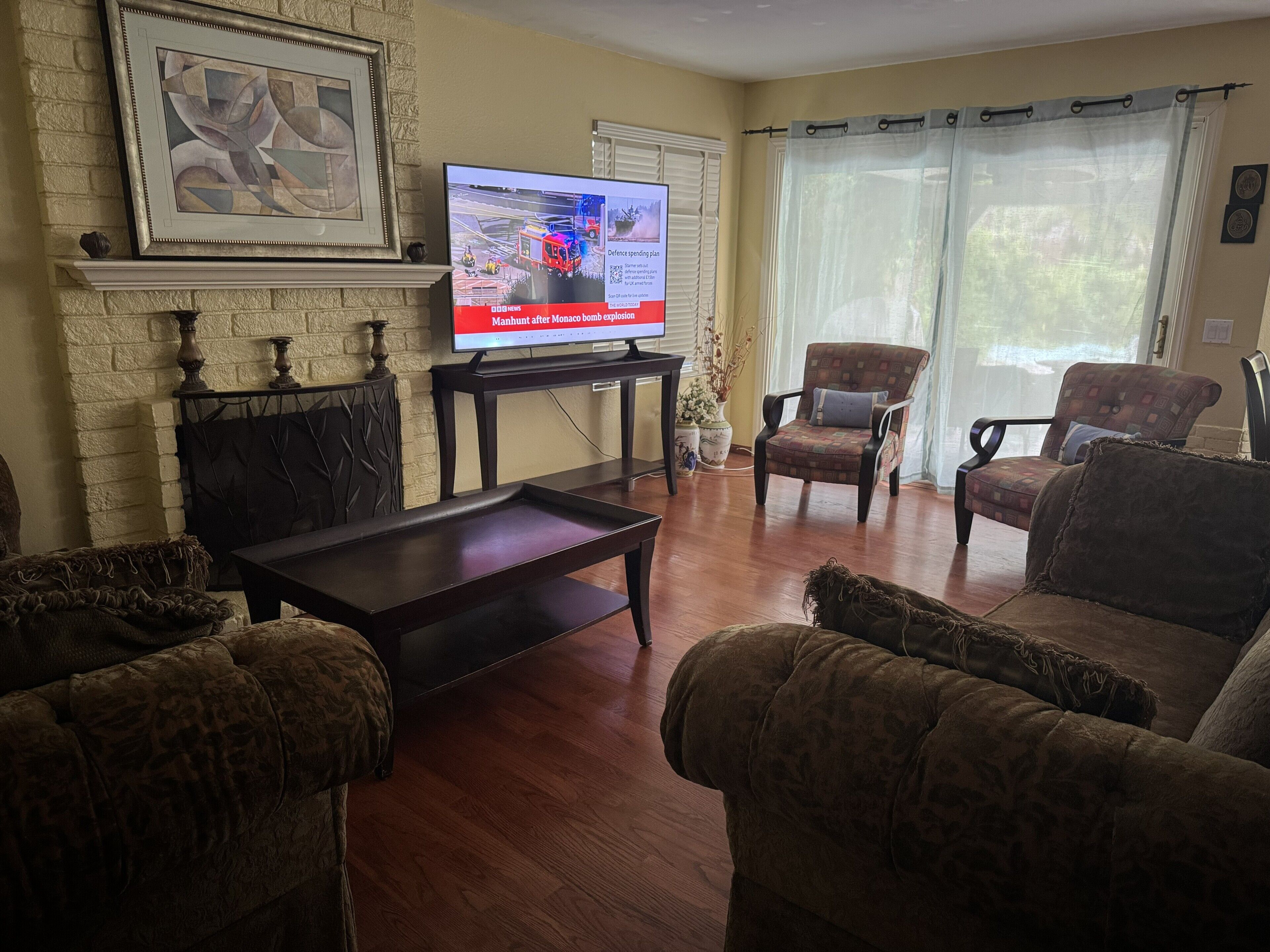 A living room with a fireplace, a television displaying news, and two armchairs.