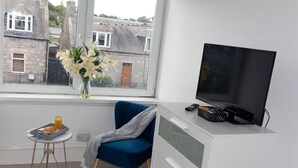 Um quarto com uma televisão sobre um móvel branco, uma cadeira azul, uma mesinha com um tabuleiro e um copo de sumo de laranja, e um vaso com flores num parapeito.