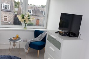 Ein Zimmer mit Fernseher auf einer weißen Kommode, einem blauen Sessel, einem kleinen Tisch mit Tablett und einem Glas Orangensaft sowie einer Vase mit Blumen auf einem Fensterbrett.