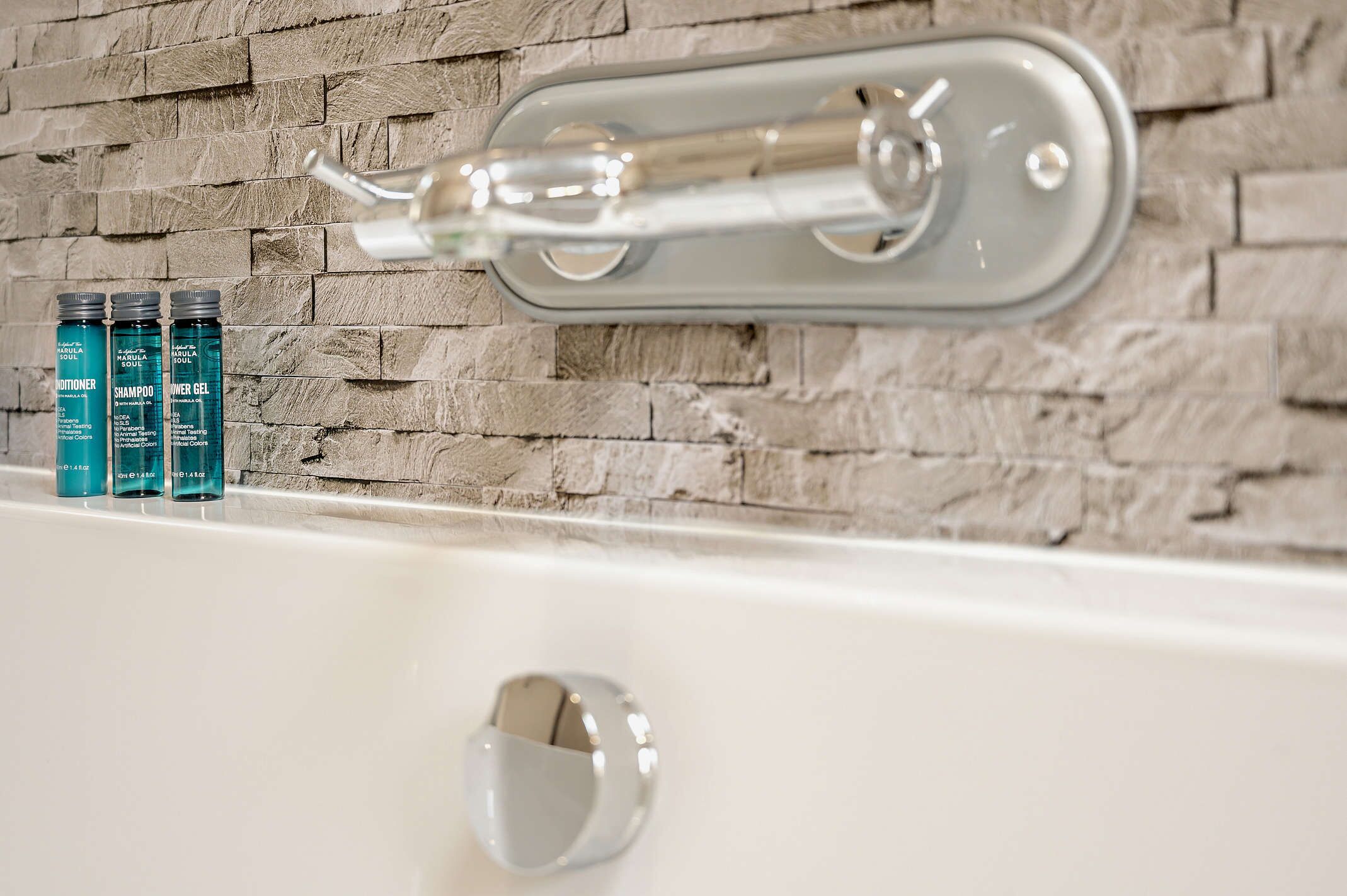 Une salle de bain avec une baignoire blanche, un robinet moderne et trois bouteilles de produits Marula Soul disposées sur le bord.