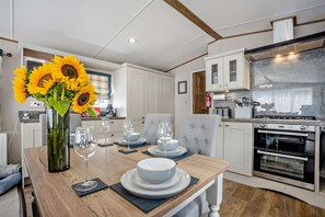 A modern kitchen with a dining area, featuring a central island with a vase of sunflowers, a built-in oven, and a coffee machine. - Kernow Lodge Luxe Seaside Cabin Wraparound Deck (Milford on Sea)