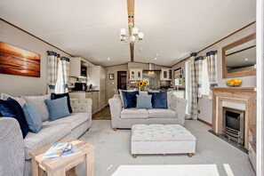 A modern living room with a sofa, ottoman, and a coffee table. There is a kitchen in the background with a stove and a dining area. - Kernow Lodge Luxe Seaside Cabin Wraparound Deck (Milford on Sea)