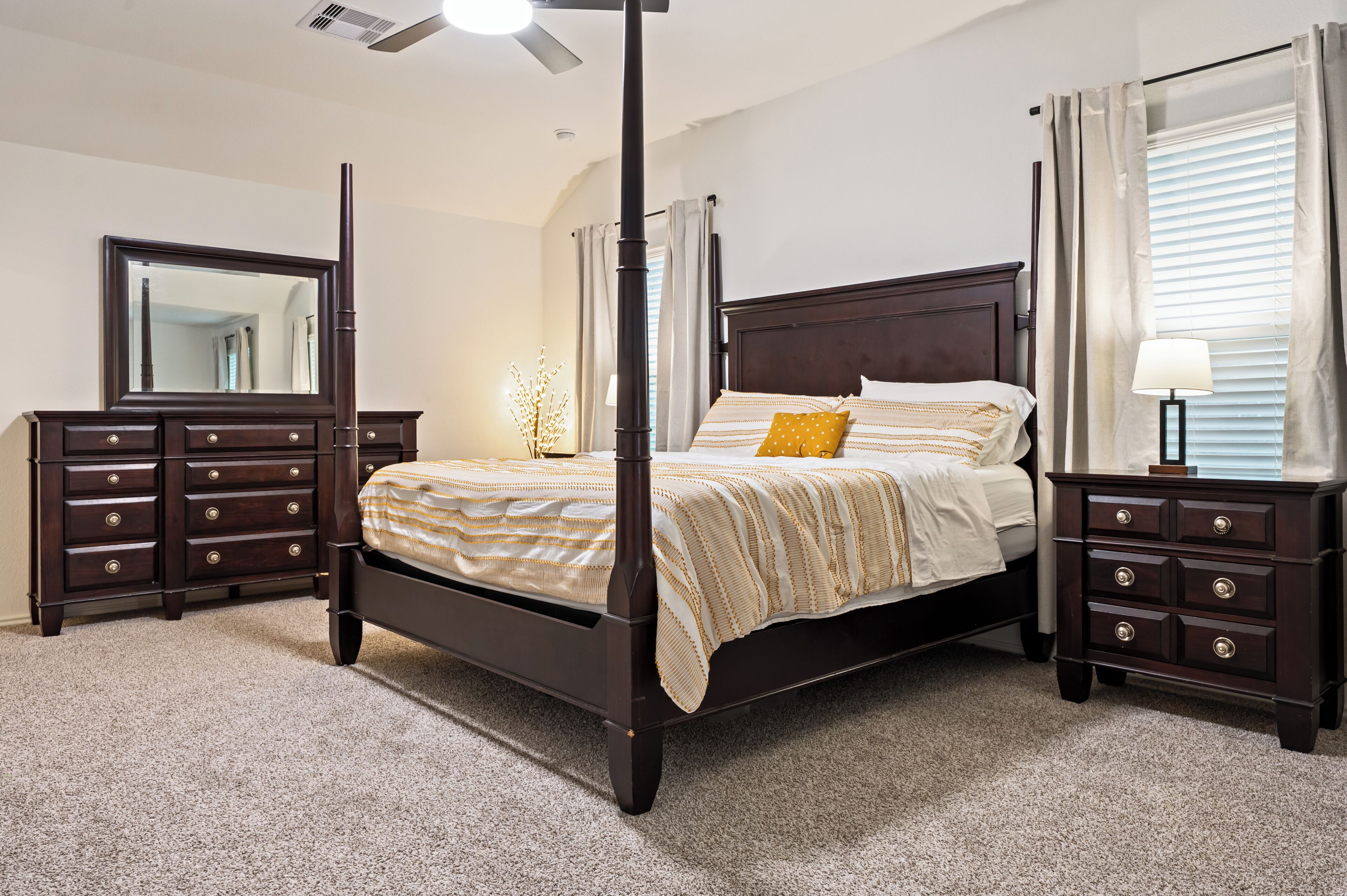 A four-poster bed with a dark finish, flanked by two nightstands with drawers, a mirror, and a floor lamp.
