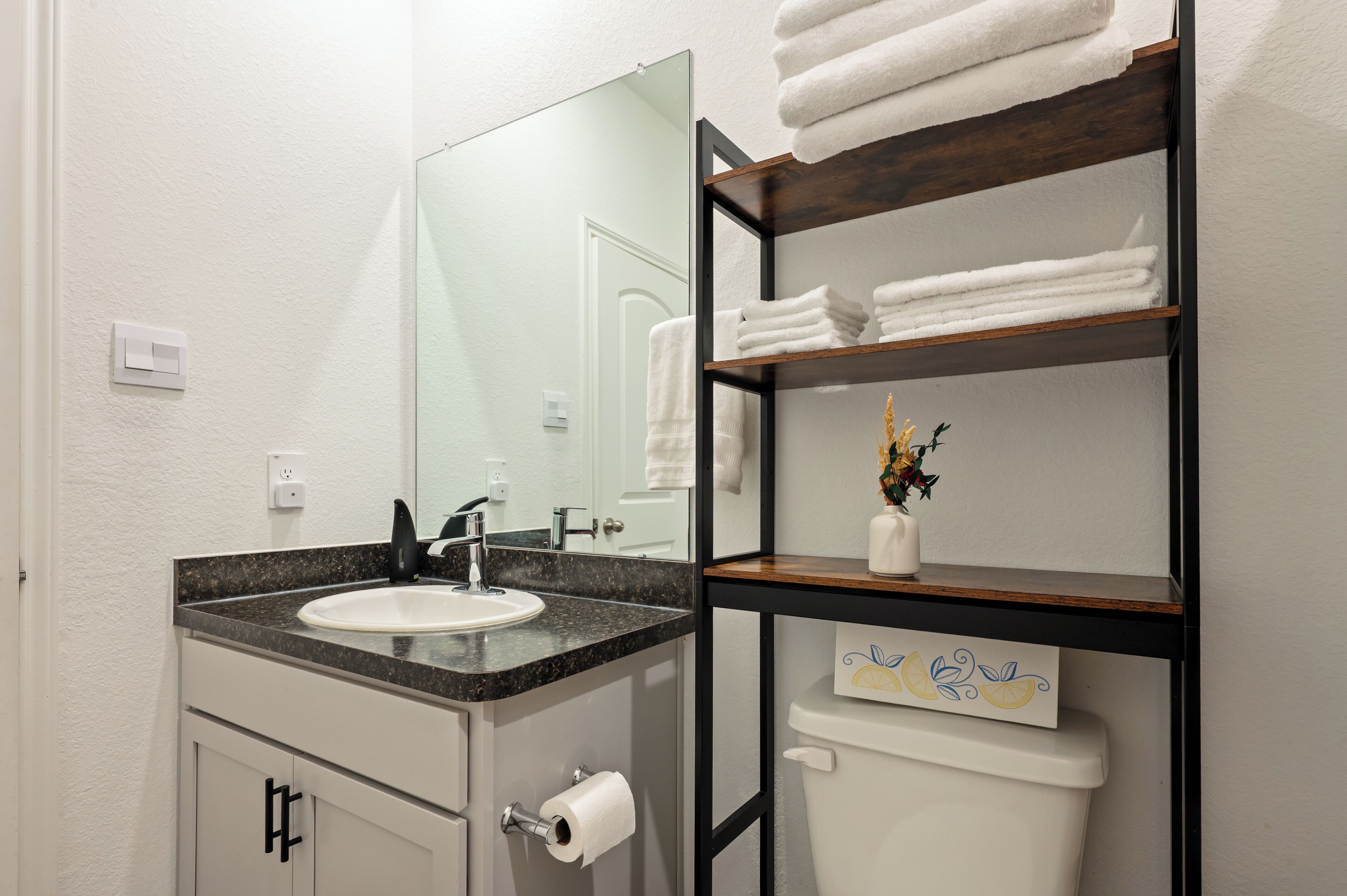 Un bagno con lavandino, specchio, porta asciugamani, porta carta igienica e ripiano con asciugamani.