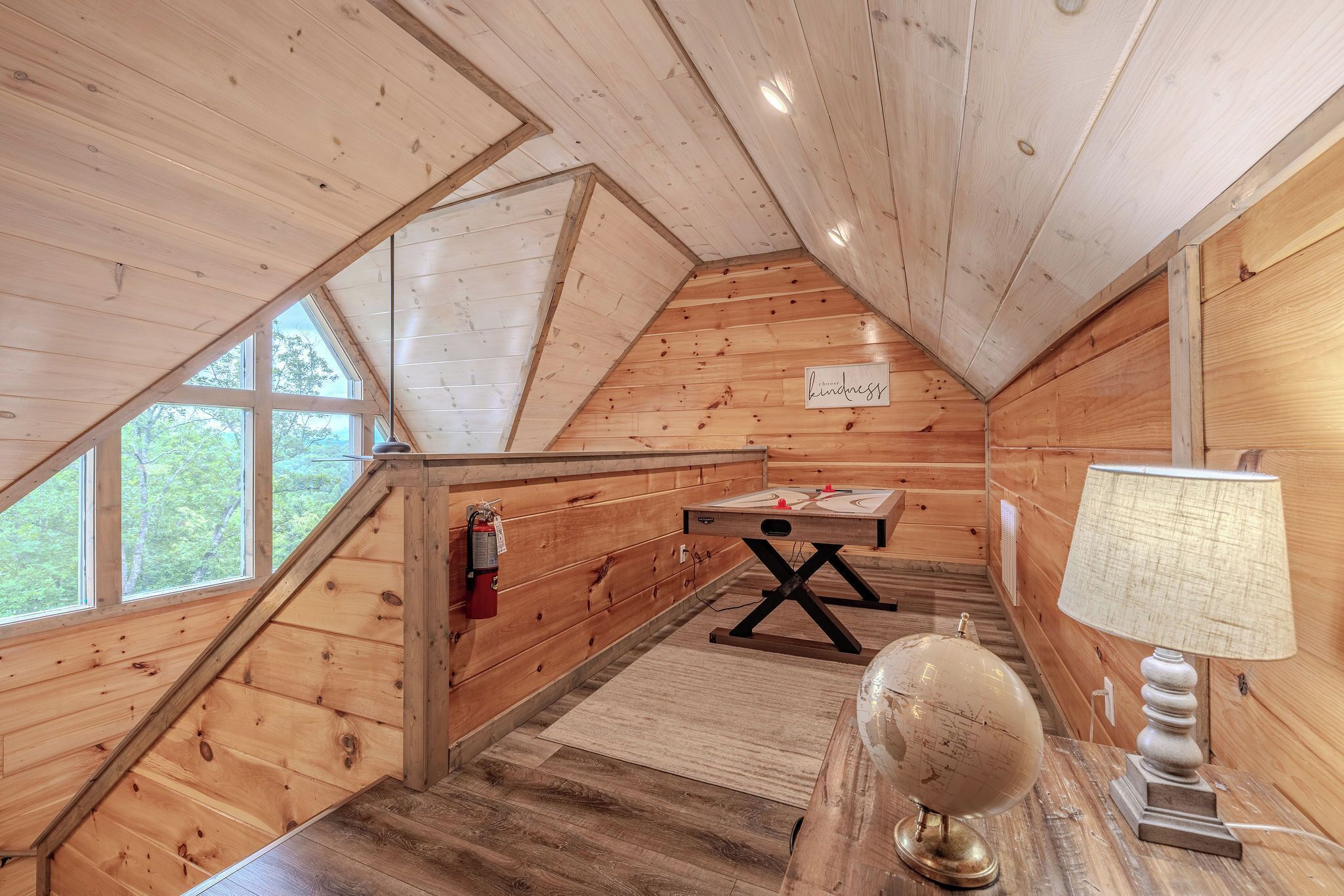 Um interior de madeira com teto inclinado, uma mesa com um globo, uma luminária e uma janela com vista para o verde.