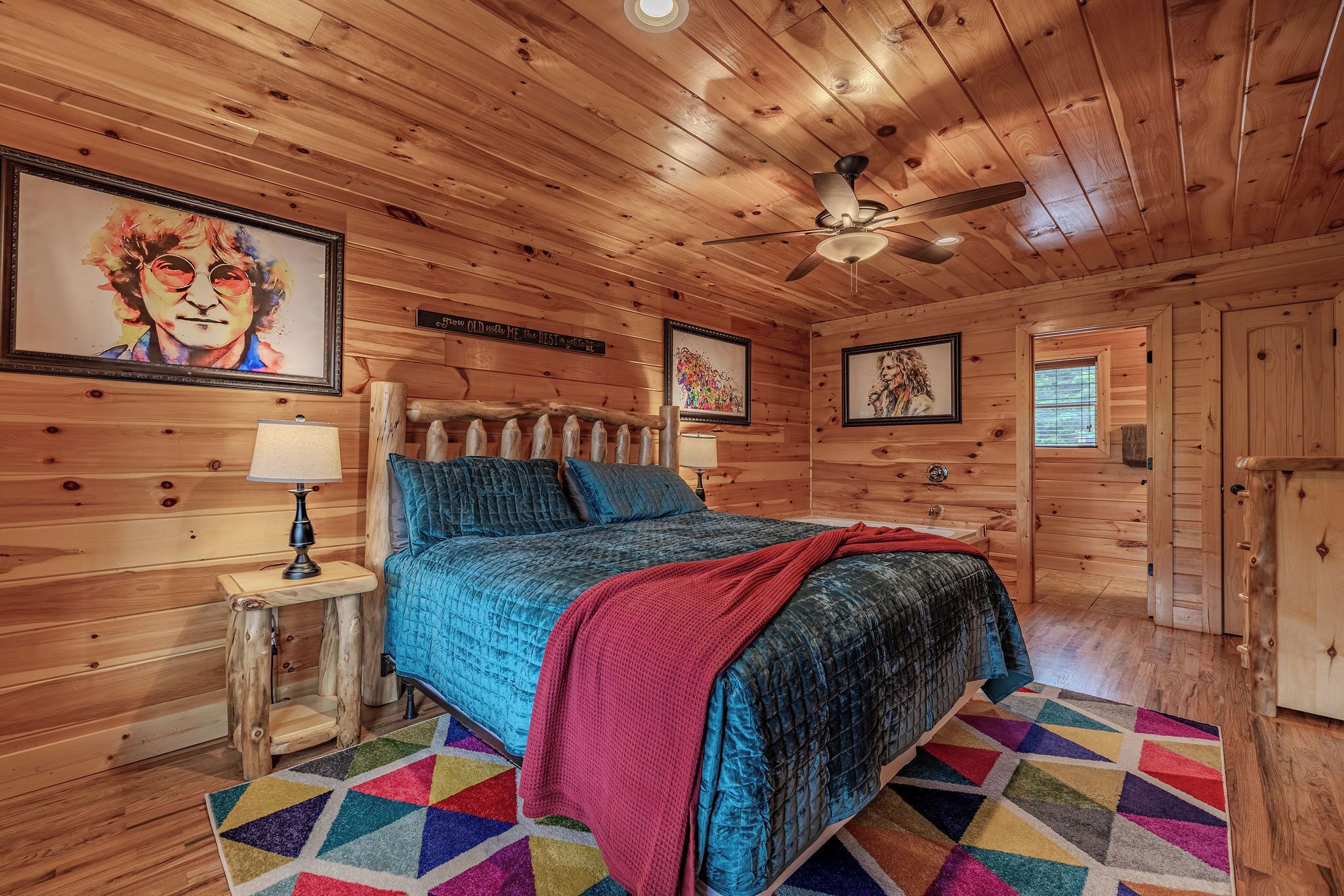 A wooden cabin bedroom with a bed, a nightstand, a lamp, a ceiling fan, and a colorful rug.