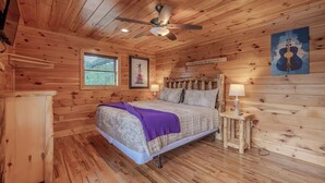 A wooden cabin bedroom with a bed, a ceiling fan, a window with blinds, a framed picture, a bedside table with a lamp, and a wall-mounted guitar artwork. - Near Downtown Gburg, Theater, Views, Trailer Parking,Dogs OK! (Gatlinburg)