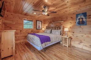 A wooden cabin bedroom with a bed, a ceiling fan, a window with blinds, a framed picture, a bedside table with a lamp, and a wall-mounted guitar artwork. - Near Downtown Gburg, Theater, Views, Trailer Parking,Dogs OK! (Gatlinburg)