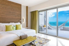Une chambre d’hôtel avec un grand lit, un banc et une porte vitrée donnant sur un balcon avec vue sur l’océan.