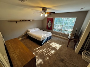 Ein Schlafzimmer mit einem Bett, einem Deckenventilator, einem Fenster mit Jalousien, einem Schreibtisch und einem an der Wand befestigten Regal.