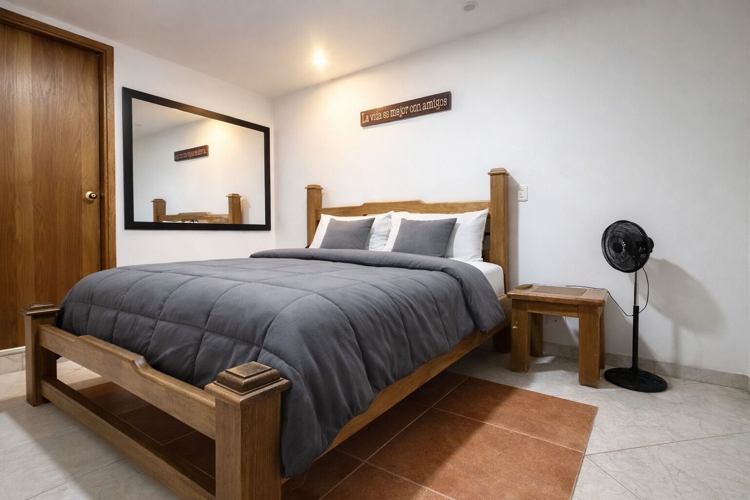 A bedroom with a wooden bed, a bedside table, a standing fan, and a mirror on the wall.