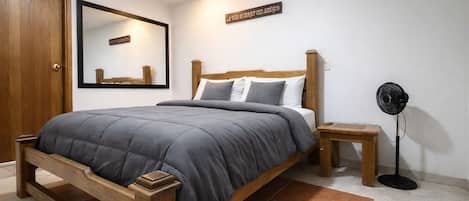 A bedroom with a wooden bed, a bedside table, a standing fan, and a mirror on the wall.